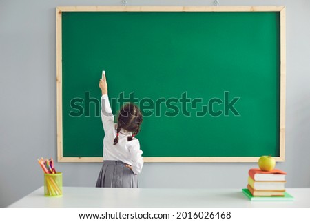 Back to school. Little schoolgirl writes on the school blackboard with chalk in the classroom.