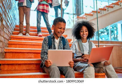 Back To School. Multiracial Pupils Of Primary School In School Hall. Children Are Ready To Study. African-American Girl And Boy Sitting On Stairs With Laptop