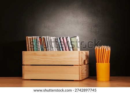 Back to school, On table there is wooden box filled with notebooks and glass with pencils against  background black school board, chalk board