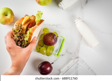 Back To School. Person Making Healthy Lunch Box With Fresh Fruit (apples, Plums, Grapes), Yogurt, Sandwich - Lettuce, Tomatoes, Cheese, Meat. White Marble Table. Copy Space Top View Female Hands