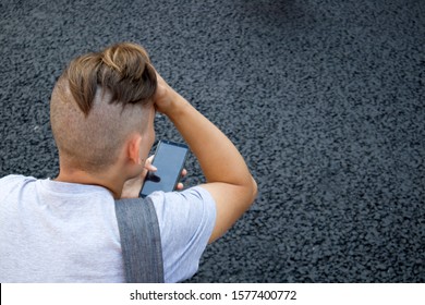 Back Of Shaved Head With Pony Tail Of Young Man With Smartphone Against Grey Asphalt