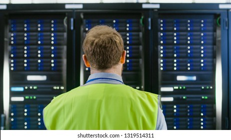 Back Shot of an IT Administrator in High Visibility Vest Walking Towards a Server Rack in Data Center Room. - Powered by Shutterstock