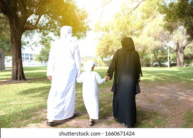 Back shot of Arab family for three. Rear view of Arabic man in dish dasha and Woman in Abaya with their son on Kandura. Walking together at a park with sunny day light