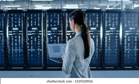 Back Shot of a Female Data Center IT Specialist Works on Laptop Computer Next to Server Rack Corridor. Running Diagnostics or Doing Maintenance Work.