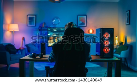 Back Shot of a Gamer Playing First-Person Shooter Online Video Game on His Powerful Personal Computer. Room and PC have Cold Colorful Neon Led Lights. Cozy Evening at Home.