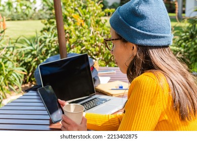 Back Shot Of A Hipster Woman Holding A Mobile Phone And A Cup With A Hot Drink With Her Laptop In The Background