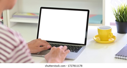 Back shot of smart man typing on white blank screen computer laptop that putting on white working desk and surrounded by coffee cup, potted plant and stationary.
