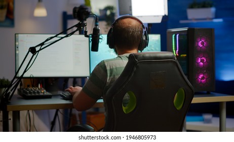 Back Shot Of Streamer Man Playing On Powerful Computer Shooter Video Game For Tournament, Talking With Multiple Players Into Headphones. Pro Video Game R Using Professional Streaming Equipment