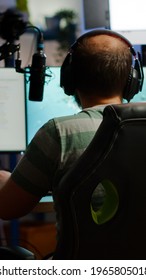 Back Shot Of Streamer Man Playing On Powerful Computer Shooter Video Game For Tournament, Talking With Multiple Players Into Headphones. Pro Video Game R Using Professional Streaming Equipment