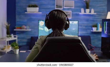 Back Shot Of Streamer Woman Playing On RGB Powerful Computer Shooter Video Game For Tournament, Talking With Multiple Players Into Headphones. Pro Video Gamer Using Professional Streaming Equipment