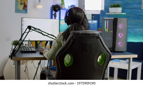 Back Shot Of Streamer Woman Playing On Powerful Computer Shooter Video Game For Tournament, Talking With Multiple Players Into Headphones. Pro Video Gamer Using Professional Streaming Equipment