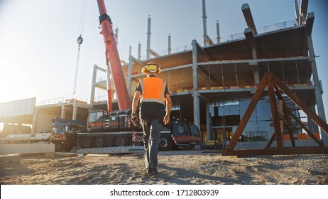 Back Shot of Worker Contractor Wearing Hard Hat and Safety Vests Walks on Industrial Building Construction Site. In the Background Crane, Skyscraper Concrete Formwork Frames