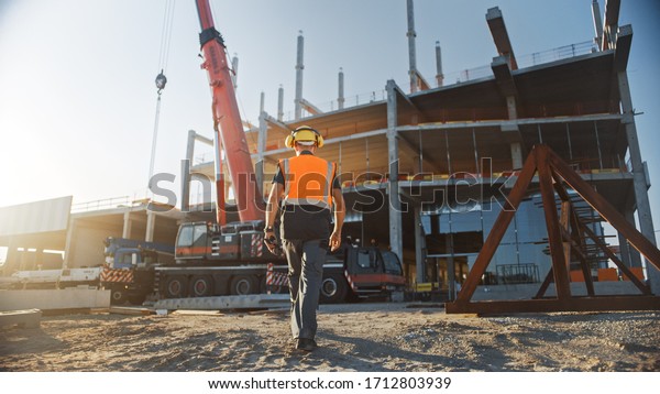 Back Shot of Worker\
Contractor Wearing Hard Hat and Safety Vests Walks on Industrial\
Building Construction Site. In the Background Crane, Skyscraper\
Concrete Formwork Frames