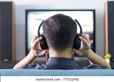 Back Side Of An Asian Man Wearing Black Heaphones In Front Of Blurry Out-of-focus Television And Home Entertainment System Background