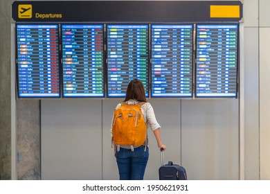 Back Side Of Asian Woman Backpacker Or Traveler With Luggage Standing Over The Flight Board For Check-in At The Flight Information Screen In Modern An Airport, Travel And Transportation Concept