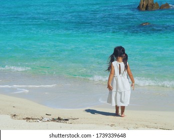 Back side little long hair girl toddler in white maxi dress standing at the beach and colorful turquoise blue sea hesitates to play the water  Sandy beach and little plastic garbage 