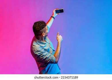 Back Side Of Man In Shirt Taking Selfie Or Broadcasting Livestream, Holding Smart Phone With Blank Screen, Showing Thumb Up. Indoor Studio Shot Isolated On Colorful Neon Light Background.