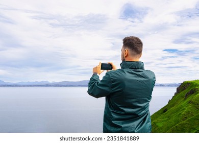 Back side shot of a young caucasian man wearing a green jacket making a photo of the landscape in the Scottish Highlands with a smartphone.