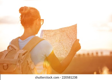 Back side of traveler girl searching right direction on map, bright orange sunset light, traveling along Europe, freedom and active lifestyle concept
