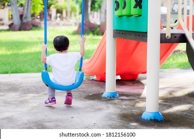 Back Side View Of Asian Toddler Boy Is Sitting On A Swing Gently. Peace Of Mind On A Winter Afternoon. Child Play In The Playground For A Relaxing Time. Baby Wearing A White Shirt Aged 2 Years Old.