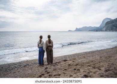 Back side view of a two young girls in casual wear stands on the seashore, best friends are looking into the distance, before sunset, dramatic cloudy weather