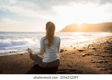 Back side view of young adult curly woman meditating in lotus yoga pose. Millennial girl practices yoga and meditations on the beach. Female yogi doing yoga at the sunset. Spiritual practice.