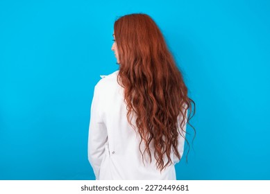 The back side view of a young red-haired doctor woman wearing medical uniform over blue studio background. Studio Shoot.
