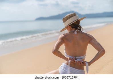 Back side woman in white bikini sitting on sand beach relaxing sunbathing.