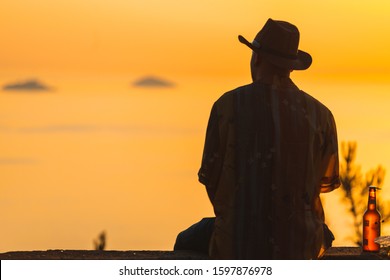 Back Silhouette View Of A Man Sitting On The Wall And Looking At Beautiful Sunset With Clear Sky In Background.