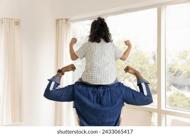 Back of strong young daddy and little kid girl sitting on fathers shoulders. Athletic dad lifting kid up, making power fist gesture, flexing hands, biceps, enjoying active spots games with daughter - Powered by Shutterstock