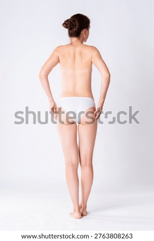 Back with a tan lines, slim topless woman in white panties on gray studio background