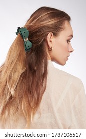 A Back Turned Blonde Girl With Golden Earrings Is Wearing A White Blouse. The Lady's Hair Is Fixed With A Hair Clip Decorated With A Green Bow With Polka Dot Print, A Golden Buckle And A Silk Ribbon. 