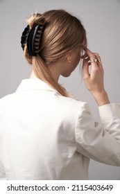 A back turned blonde girl with golden earrings and rings is wearing a white blouse. The lady's hair is fixed with semicircular black hair clip. The woman is posing on the gray backdrop.