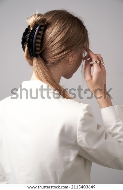 A back\
turned blonde girl with golden earrings and rings is wearing a\
white blouse. The lady\'s hair is fixed with semicircular black hair\
clip. The woman is posing on the gray\
backdrop.