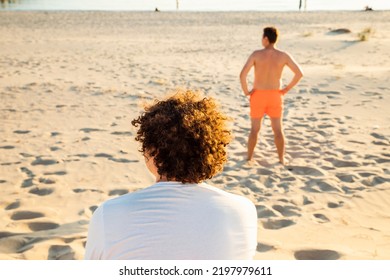 Back Turned Look Away Two Young Men On Sandy Summer Beach. Curly Hair Man Is Sitting, Another Man Is Lifeguard In Red Swim Shorts. 