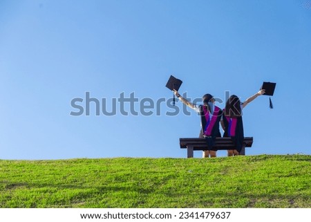 Back of Two friends in graduation caps and gowns sitting on the bench look blue sky and think future