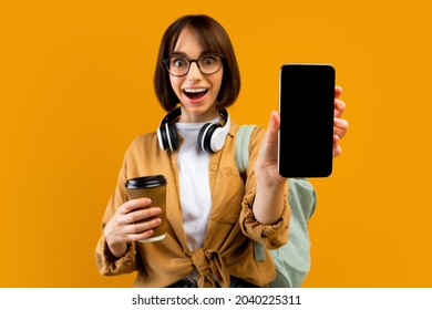 Back To University Concept. Excited Female Student Showing Smartphone With Empty Screen, Posing With Backpack, Headphones And Coffee On Yellow Studio Background, Mockup For App Or Website Design