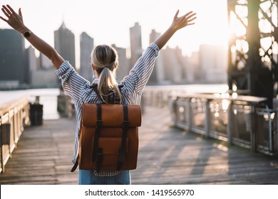 Back view of accomplished tourist woman with trendy backpack and raised hands celebrating win with trip to American city - New York, female traveller complete triumph and win goal during journey