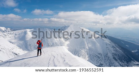 Back view of active man ski touring at mountains background at sunny winter day. Ski mountaineer with red jacket walking up along a steep snowy ridge with the skis in the backpack