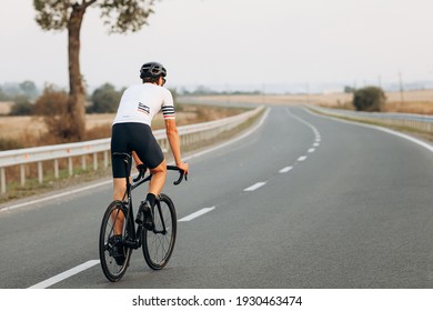 Back View Of Active Man In White T-shirt, Black Shorts And Protective Helmet Riding Bike On Fresh Air. Concept Of Competition And Regular Training.