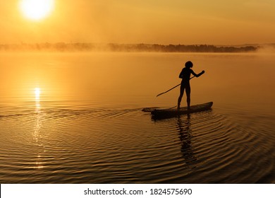 Back view of active young man using long paddle for swimming on sup board. Light waves on water surface. Heavy fog around lake.