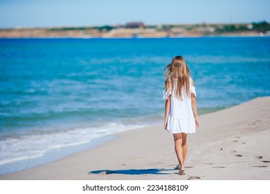 Back view of adorable little girl with long hair in white dress walking on tropical beach vacation