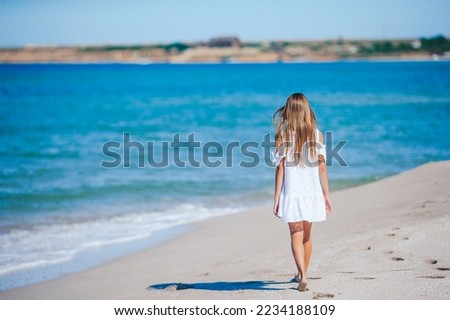 Back view of adorable little girl with long hair in white dress walking on tropical beach vacation