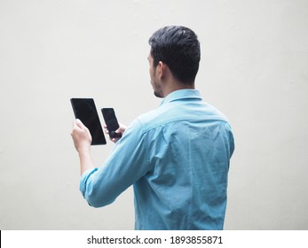 Back View Of Adult Asian Man Wearing Blue Shirt Holding And Looking To Mobile Phone And Tablet. Isolated On White