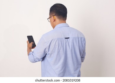 Back View Of Adult Asian Man Wearing Bright Blue Shirt Looking To Mobile Phone Screen. Isolated On White