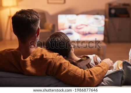 Back view of adult couple watching TV at home while sitting on sofa lit by warm cozy light, copy space