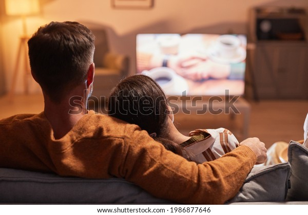 Back view of adult couple\
watching TV at home while sitting on sofa lit by warm cozy light,\
copy space
