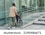 Back view of adult man assisting partner in wheelchair going up accessibility ramp in city, copy space