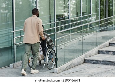 Back view of adult man assisting partner in wheelchair going up accessibility ramp in city, copy space - Powered by Shutterstock