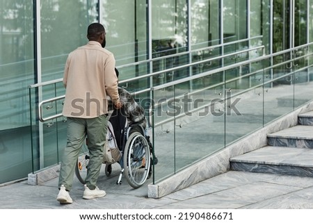 Back view of adult man assisting partner in wheelchair going up accessibility ramp in city, copy space [[stock_photo]] © 